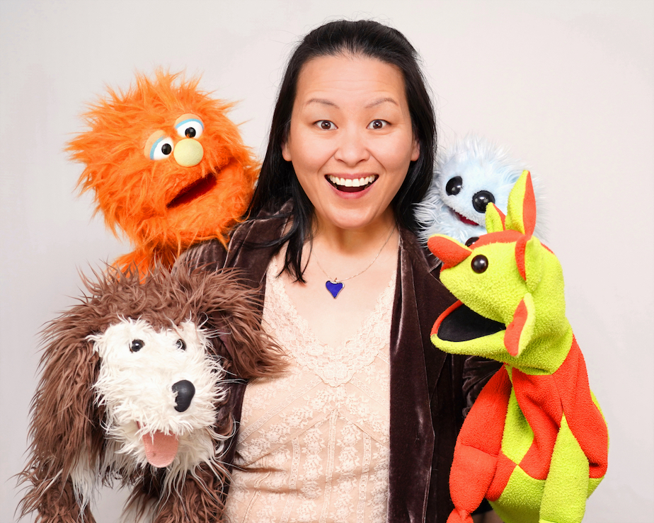 image shows leigh baltzer with an excited smile, surrounded by four different puppets